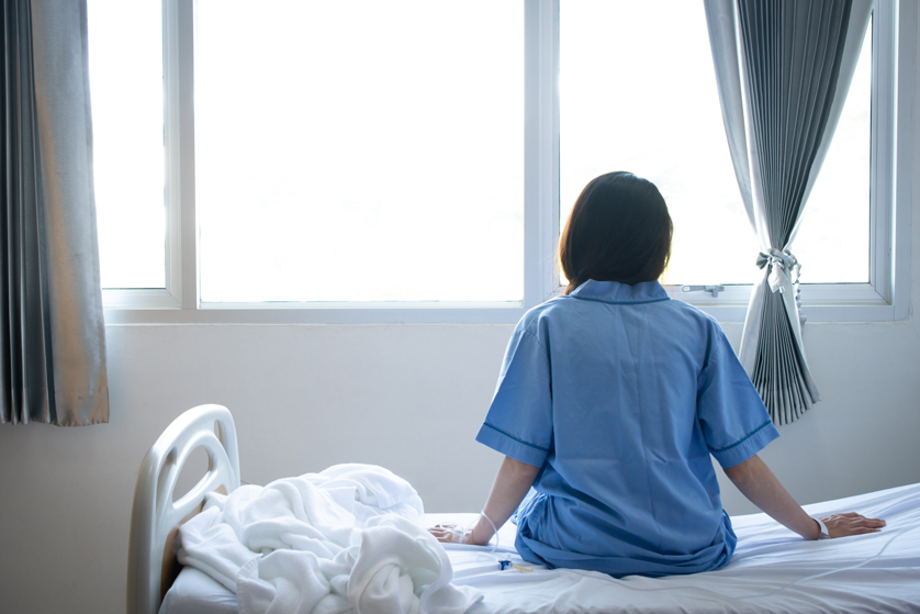 Woman misdiagnosed with cancer sits on the edge of a hospital bed staring out the window.